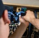 A technician wearing a dark cap is using a red-handled screwdriver to work on a complex piece of electronic or electromechanical equipment. The equipment appears to be an older, rack-mounted or cabinet-style unit with multiple circuit boards, wiring, tubes, and labeled components. The work appears to be maintenance, repair, or assembly of specialized technical hardware.