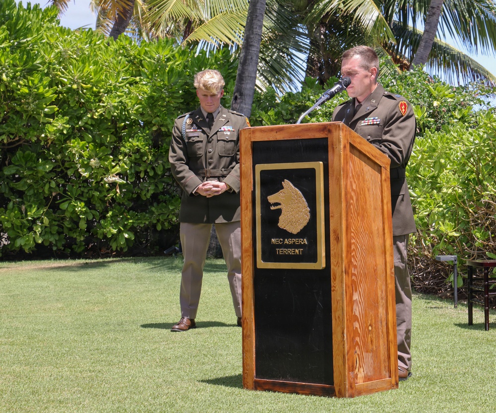 Two U.S. Army officers in Class A dress uniforms are participating in an outdoor military ceremony, likely in Hawaii based on the tropical vegetation. One officer stands at a wooden podium bearing a wolf/dog head insignia and the Latin motto 'NEC ASPERA TERRENT' (meaning 'Nor do difficulties deter'), speaking into a microphone. The second officer stands at attention in the background with hands clasped.