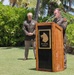 Two U.S. Army officers in Class A dress uniforms are participating in an outdoor military ceremony, likely in Hawaii based on the tropical vegetation. One officer stands at a wooden podium bearing a wolf/dog head insignia and the Latin motto 'NEC ASPERA TERRENT' (meaning 'Nor do difficulties deter'), speaking into a microphone. The second officer stands at attention in the background with hands clasped.