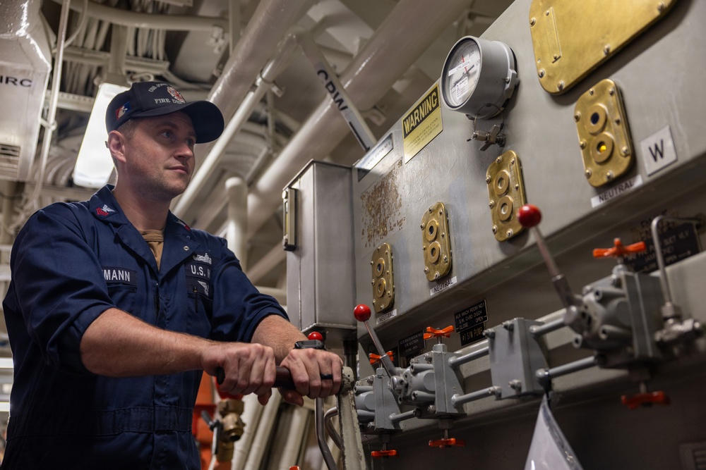 A U.S. military servicemember identified by name tape as 'MANN' is working at an industrial control panel aboard what appears to be a naval vessel. He is wearing dark navy coveralls with 'U.S.' branch tape and a cap with 'FIRE DEPT' visible. The setting is a ship's mechanical or engineering space with pipes, valves, gauges, and brass fittings.