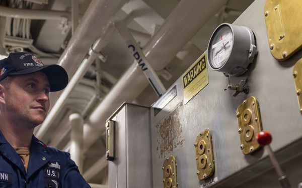 A U.S. military servicemember identified by name tape as 'MANN' is working at an industrial control panel aboard what appears to be a naval vessel. He is wearing dark navy coveralls with 'U.S.' branch tape and a cap with 'FIRE DEPT' visible. The setting is a ship's mechanical or engineering space with pipes, valves, gauges, and brass fittings.