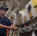 A U.S. military servicemember identified by name tape as 'MANN' is working at an industrial control panel aboard what appears to be a naval vessel. He is wearing dark navy coveralls with 'U.S.' branch tape and a cap with 'FIRE DEPT' visible. The setting is a ship's mechanical or engineering space with pipes, valves, gauges, and brass fittings.