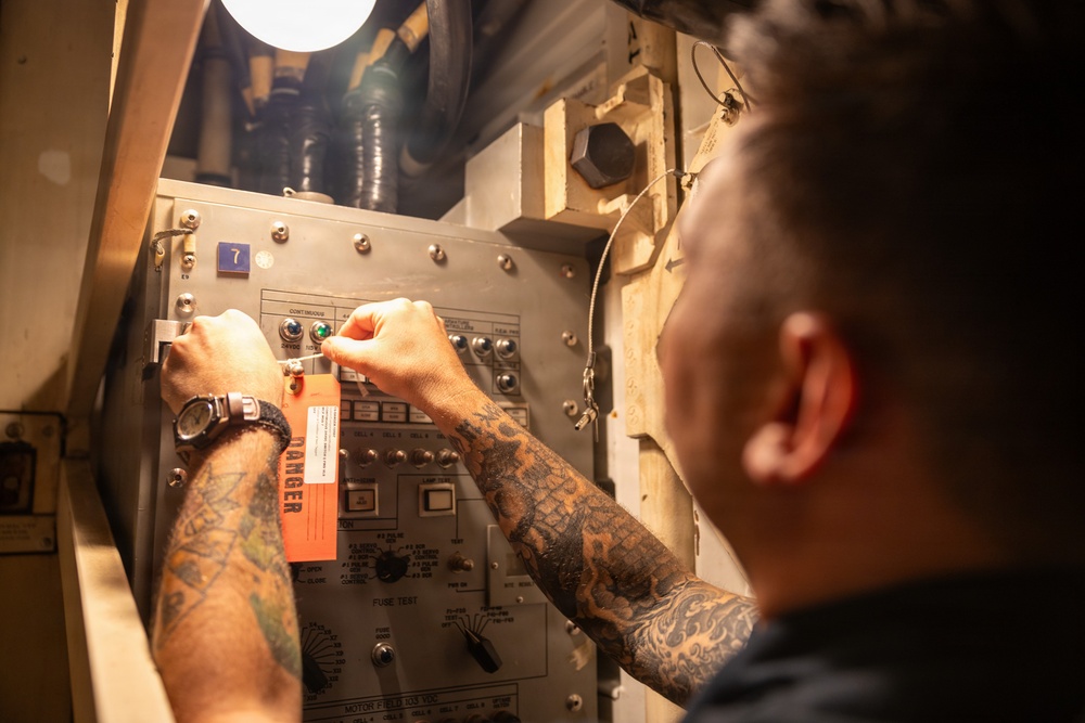 A heavily tattooed individual is working on an industrial or military-grade electrical control panel in a confined, dimly lit space. The person is manipulating switches or controls on the panel, which has a prominent orange 'DANGER' tag attached to it. The setting appears to be aboard a naval vessel or similar industrial environment based on the panel design and surrounding infrastructure.