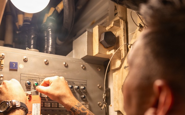 A heavily tattooed individual is working on an industrial or military-grade electrical control panel in a confined, dimly lit space. The person is manipulating switches or controls on the panel, which has a prominent orange 'DANGER' tag attached to it. The setting appears to be aboard a naval vessel or similar industrial environment based on the panel design and surrounding infrastructure.