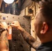 A heavily tattooed individual is working on an industrial or military-grade electrical control panel in a confined, dimly lit space. The person is manipulating switches or controls on the panel, which has a prominent orange 'DANGER' tag attached to it. The setting appears to be aboard a naval vessel or similar industrial environment based on the panel design and surrounding infrastructure.