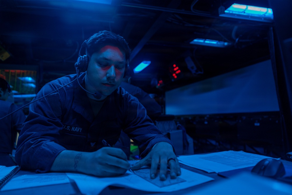 A U.S. Navy sailor is working at a dimly lit station aboard what appears to be a naval vessel, writing on documents while wearing a communications headset. The environment is illuminated predominantly by blue lighting, characteristic of a ship's combat information center or bridge during low-light operations. Multiple electronic displays and indicator lights are visible in the background.