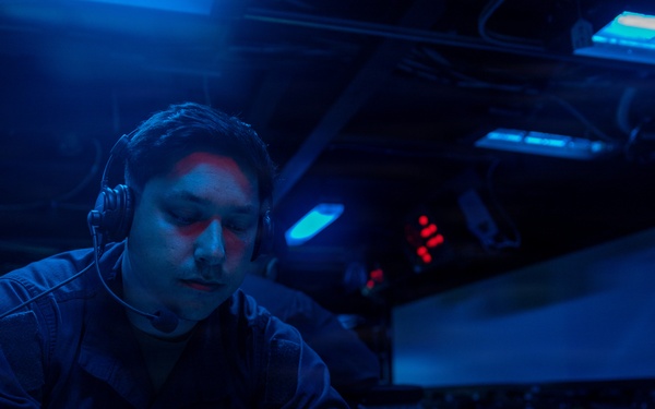 A U.S. Navy sailor is working at a dimly lit station aboard what appears to be a naval vessel, writing on documents while wearing a communications headset. The environment is illuminated predominantly by blue lighting, characteristic of a ship's combat information center or bridge during low-light operations. Multiple electronic displays and indicator lights are visible in the background.