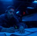 A U.S. Navy sailor is working at a dimly lit station aboard what appears to be a naval vessel, writing on documents while wearing a communications headset. The environment is illuminated predominantly by blue lighting, characteristic of a ship's combat information center or bridge during low-light operations. Multiple electronic displays and indicator lights are visible in the background.