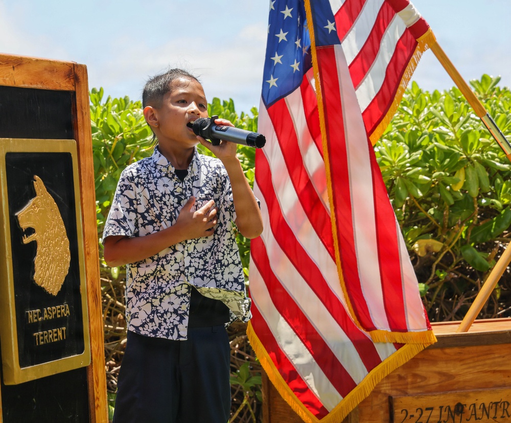 A young child is singing or performing into a handheld microphone at what appears to be a military ceremony, with one hand placed over their chest. The setting includes an American flag with gold fringe, a wooden podium marked '2-27 INFANTR[Y]', and a plaque bearing a wolf emblem and the Latin motto 'NEC ASPERA TERRENT' (meaning 'Difficulties do not deter'). The event appears to be an outdoor military unit ceremony, likely at a U.S. Army installation.