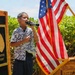 A young child is singing or performing into a handheld microphone at what appears to be a military ceremony, with one hand placed over their chest. The setting includes an American flag with gold fringe, a wooden podium marked '2-27 INFANTR[Y]', and a plaque bearing a wolf emblem and the Latin motto 'NEC ASPERA TERRENT' (meaning 'Difficulties do not deter'). The event appears to be an outdoor military unit ceremony, likely at a U.S. Army installation.