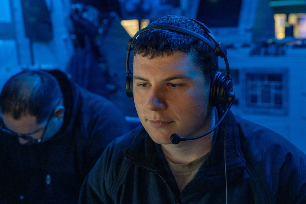 A young man wearing a black headset with a boom microphone is seated in a dimly lit environment illuminated by blue ambient lighting. He appears focused, looking downward or slightly to the side, suggesting active communication or monitoring. A second individual wearing glasses is partially visible in the background to the left.