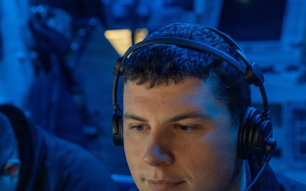A young man wearing a black headset with a boom microphone is seated in a dimly lit environment illuminated by blue ambient lighting. He appears focused, looking downward or slightly to the side, suggesting active communication or monitoring. A second individual wearing glasses is partially visible in the background to the left.