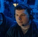A young man wearing a black headset with a boom microphone is seated in a dimly lit environment illuminated by blue ambient lighting. He appears focused, looking downward or slightly to the side, suggesting active communication or monitoring. A second individual wearing glasses is partially visible in the background to the left.