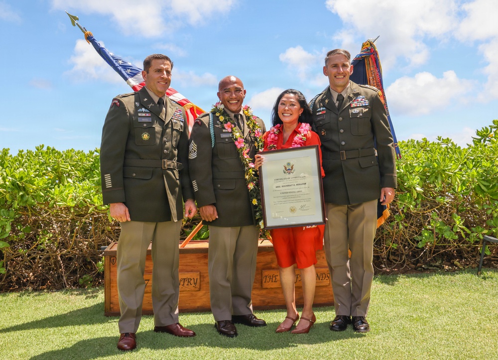 A military ceremony photograph showing four individuals posing outdoors in what appears to be Hawaii, indicated by the floral leis worn by two of the subjects. Three individuals are wearing U.S. Army dress green uniforms, while one woman in a red outfit holds a framed certificate. The setting includes U.S. and military unit flags, a wooden display box, and lush green hedges under a partly cloudy sky.