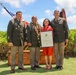 A military ceremony photograph showing four individuals posing outdoors in what appears to be Hawaii, indicated by the floral leis worn by two of the subjects. Three individuals are wearing U.S. Army dress green uniforms, while one woman in a red outfit holds a framed certificate. The setting includes U.S. and military unit flags, a wooden display box, and lush green hedges under a partly cloudy sky.