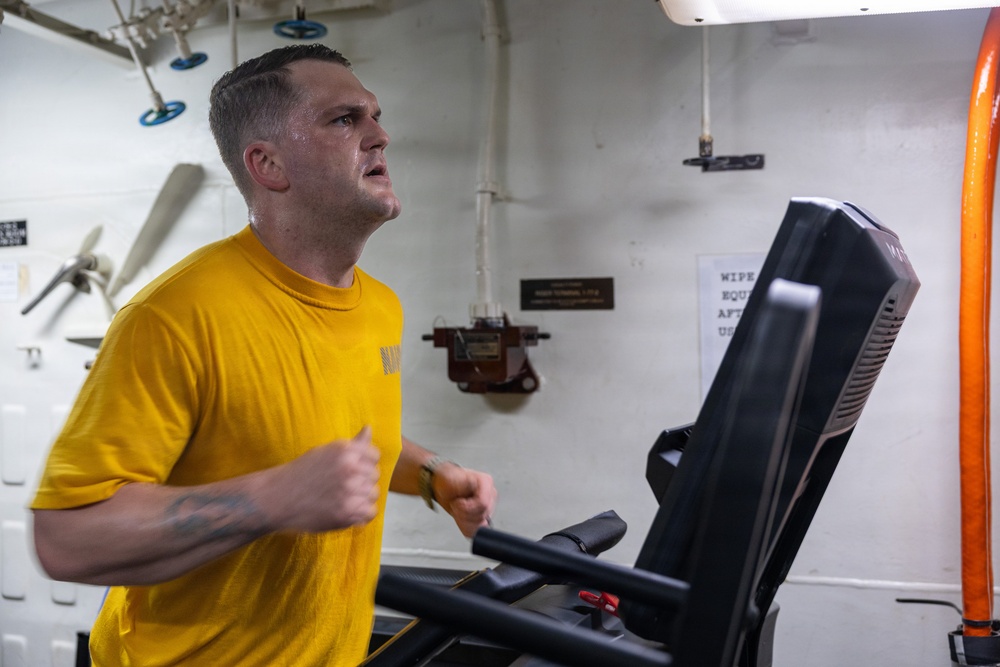 A man in a yellow 'NAVY' t-shirt is running intensely on a treadmill in what appears to be a shipboard fitness facility. He is visibly sweating and exerting significant effort, with clenched fists and an open mouth. The setting features white metal walls with pipes, ventilation equipment, and a posted sign reminding users to wipe down equipment after use.