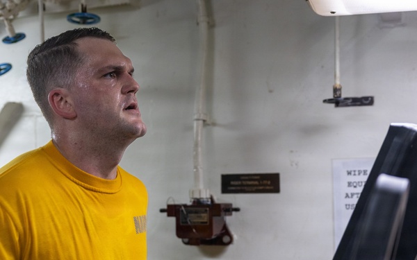 A man in a yellow 'NAVY' t-shirt is running intensely on a treadmill in what appears to be a shipboard fitness facility. He is visibly sweating and exerting significant effort, with clenched fists and an open mouth. The setting features white metal walls with pipes, ventilation equipment, and a posted sign reminding users to wipe down equipment after use.