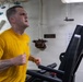 A man in a yellow 'NAVY' t-shirt is running intensely on a treadmill in what appears to be a shipboard fitness facility. He is visibly sweating and exerting significant effort, with clenched fists and an open mouth. The setting features white metal walls with pipes, ventilation equipment, and a posted sign reminding users to wipe down equipment after use.