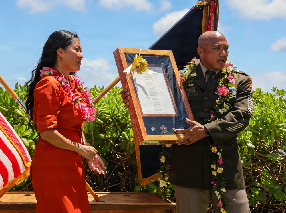 A military ceremony appears to be taking place outdoors in what looks like Hawaii, based on the tropical vegetation and traditional flower leis worn by both participants. A U.S. Army soldier in dress uniform and a woman in a red dress are jointly presenting or receiving a framed document or plaque decorated with a yellow bow. The setting includes an American flag and a military unit guidon or color in the background.