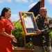 A military ceremony appears to be taking place outdoors in what looks like Hawaii, based on the tropical vegetation and traditional flower leis worn by both participants. A U.S. Army soldier in dress uniform and a woman in a red dress are jointly presenting or receiving a framed document or plaque decorated with a yellow bow. The setting includes an American flag and a military unit guidon or color in the background.