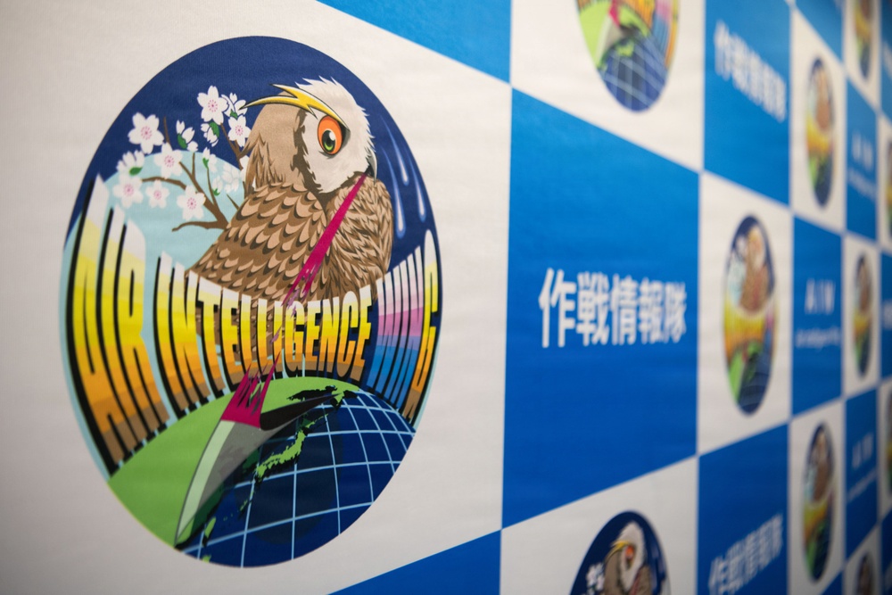 The image shows a display banner or backdrop featuring the emblem of an 'Air Intelligence Wing,' likely associated with a Japanese military or defense organization. The central logo depicts an owl perched on a globe with cherry blossoms, accompanied by Japanese text reading '作戦情報隊' (Operations Intelligence Squadron). The background follows a blue and white checkered pattern with multiple smaller circular emblems visible.