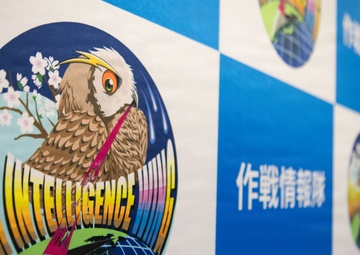 The image shows a display banner or backdrop featuring the emblem of an 'Air Intelligence Wing,' likely associated with a Japanese military or defense organization. The central logo depicts an owl perched on a globe with cherry blossoms, accompanied by Japanese text reading '作戦情報隊' (Operations Intelligence Squadron). The background follows a blue and white checkered pattern with multiple smaller circular emblems visible.