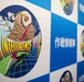 The image shows a display banner or backdrop featuring the emblem of an 'Air Intelligence Wing,' likely associated with a Japanese military or defense organization. The central logo depicts an owl perched on a globe with cherry blossoms, accompanied by Japanese text reading '作戦情報隊' (Operations Intelligence Squadron). The background follows a blue and white checkered pattern with multiple smaller circular emblems visible.