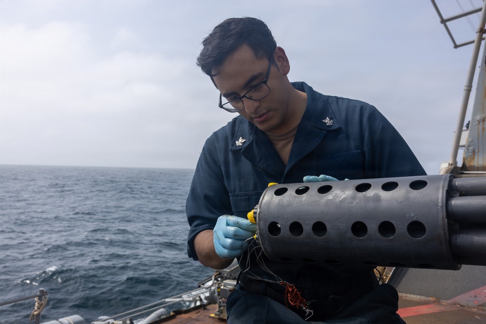 A U.S. Navy sailor wearing dark blue coveralls and blue nitrile gloves is working on a cylindrical metal device with perforated holes on the deck of a naval vessel. The sailor, identifiable by Petty Officer Third Class collar insignia, appears to be handling a yellow component connected to wiring on what appears to be a sonobuoy launcher or similar naval equipment. The setting is an overcast open ocean environment aboard a warship.