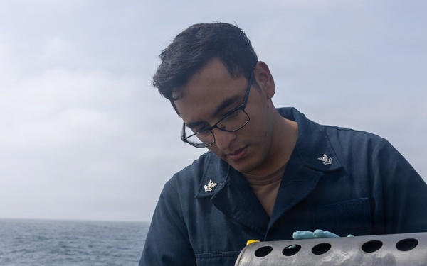 A U.S. Navy sailor wearing dark blue coveralls and blue nitrile gloves is working on a cylindrical metal device with perforated holes on the deck of a naval vessel. The sailor, identifiable by Petty Officer Third Class collar insignia, appears to be handling a yellow component connected to wiring on what appears to be a sonobuoy launcher or similar naval equipment. The setting is an overcast open ocean environment aboard a warship.