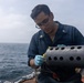 A U.S. Navy sailor wearing dark blue coveralls and blue nitrile gloves is working on a cylindrical metal device with perforated holes on the deck of a naval vessel. The sailor, identifiable by Petty Officer Third Class collar insignia, appears to be handling a yellow component connected to wiring on what appears to be a sonobuoy launcher or similar naval equipment. The setting is an overcast open ocean environment aboard a warship.
