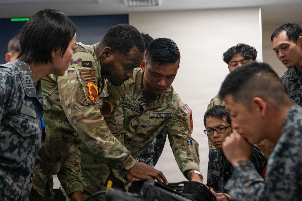 The image shows a group of military personnel from multiple nations gathered around a table in what appears to be an indoor briefing or training environment. Two U.S. Space Force and U.S. Army soldiers in OCP camouflage uniforms are leaning over and pointing at equipment or documents on the table, while personnel in what appear to be Asian military uniforms observe closely. The scene suggests a multinational military training exercise or joint operations briefing.