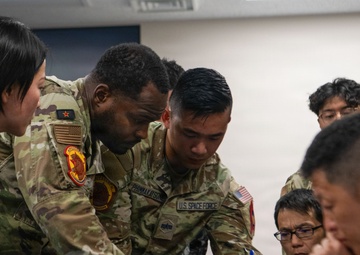 The image shows a group of military personnel from multiple nations gathered around a table in what appears to be an indoor briefing or training environment. Two U.S. Space Force and U.S. Army soldiers in OCP camouflage uniforms are leaning over and pointing at equipment or documents on the table, while personnel in what appear to be Asian military uniforms observe closely. The scene suggests a multinational military training exercise or joint operations briefing.
