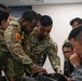 The image shows a group of military personnel from multiple nations gathered around a table in what appears to be an indoor briefing or training environment. Two U.S. Space Force and U.S. Army soldiers in OCP camouflage uniforms are leaning over and pointing at equipment or documents on the table, while personnel in what appear to be Asian military uniforms observe closely. The scene suggests a multinational military training exercise or joint operations briefing.