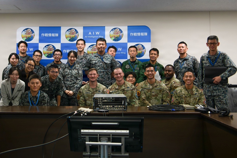 A group photo of military personnel from what appears to be a joint US-Japanese military gathering at the Air Intelligence Wing (AIW). The backdrop displays the AIW logo alongside Japanese text reading '作戦情報隊' (Air Intelligence Squadron/Unit). Personnel are wearing a mix of Japanese Self-Defense Force camouflage uniforms and US Army/Air Force uniforms.