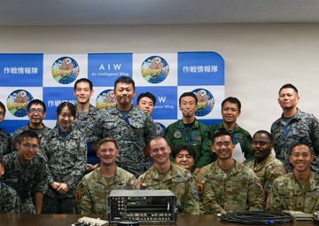A group photo of military personnel from what appears to be a joint US-Japanese military gathering at the Air Intelligence Wing (AIW). The backdrop displays the AIW logo alongside Japanese text reading '作戦情報隊' (Air Intelligence Squadron/Unit). Personnel are wearing a mix of Japanese Self-Defense Force camouflage uniforms and US Army/Air Force uniforms.