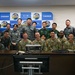 A group photo of military personnel from what appears to be a joint US-Japanese military gathering at the Air Intelligence Wing (AIW). The backdrop displays the AIW logo alongside Japanese text reading '作戦情報隊' (Air Intelligence Squadron/Unit). Personnel are wearing a mix of Japanese Self-Defense Force camouflage uniforms and US Army/Air Force uniforms.