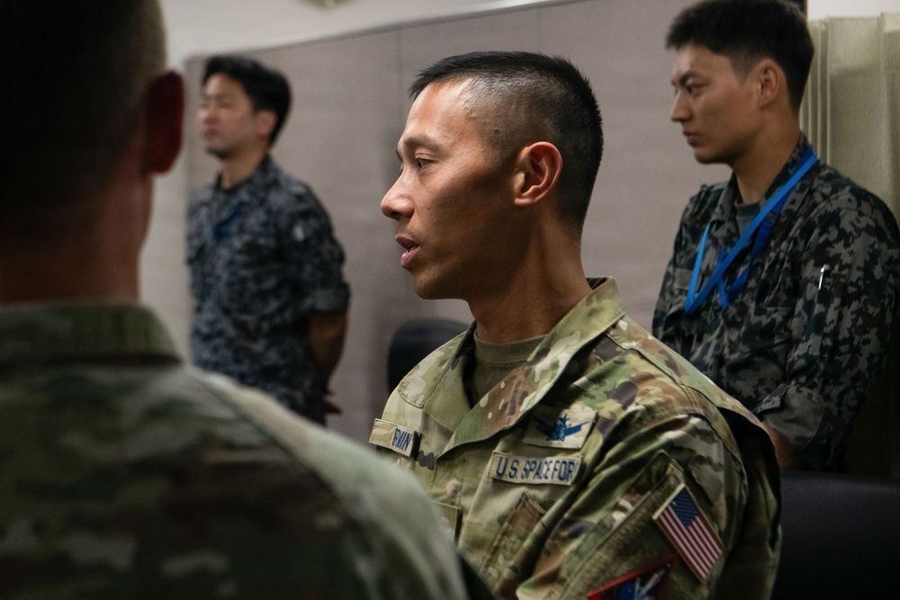 A U.S. Space Force service member in MultiCam/OCP uniform is engaged in conversation with another military personnel in the foreground. In the background, two individuals wearing what appears to be Japanese Maritime Self-Defense Force (JMSDF) digital camouflage uniforms are visible. The setting appears to be an indoor military facility, suggesting a joint multinational military engagement or exercise.