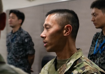 A U.S. Space Force service member in MultiCam/OCP uniform is engaged in conversation with another military personnel in the foreground. In the background, two individuals wearing what appears to be Japanese Maritime Self-Defense Force (JMSDF) digital camouflage uniforms are visible. The setting appears to be an indoor military facility, suggesting a joint multinational military engagement or exercise.
