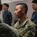 A U.S. Space Force service member in MultiCam/OCP uniform is engaged in conversation with another military personnel in the foreground. In the background, two individuals wearing what appears to be Japanese Maritime Self-Defense Force (JMSDF) digital camouflage uniforms are visible. The setting appears to be an indoor military facility, suggesting a joint multinational military engagement or exercise.