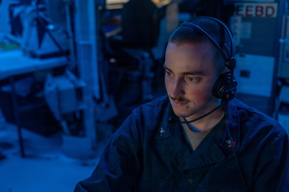 A young male military personnel, likely a U.S. Navy sailor, is seated at a workstation in what appears to be a combat information center or operations room aboard a naval vessel. He is wearing a dark navy coverall uniform and a communications headset, focused intently on a screen in front of him. The environment is illuminated with blue ambient lighting, characteristic of shipboard operations spaces.