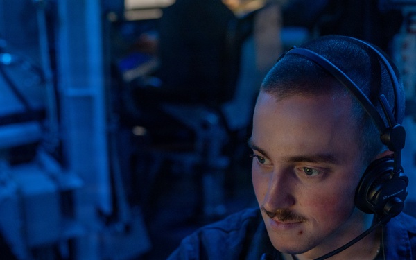 A young male military personnel, likely a U.S. Navy sailor, is seated at a workstation in what appears to be a combat information center or operations room aboard a naval vessel. He is wearing a dark navy coverall uniform and a communications headset, focused intently on a screen in front of him. The environment is illuminated with blue ambient lighting, characteristic of shipboard operations spaces.