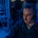 A young male military personnel, likely a U.S. Navy sailor, is seated at a workstation in what appears to be a combat information center or operations room aboard a naval vessel. He is wearing a dark navy coverall uniform and a communications headset, focused intently on a screen in front of him. The environment is illuminated with blue ambient lighting, characteristic of shipboard operations spaces.