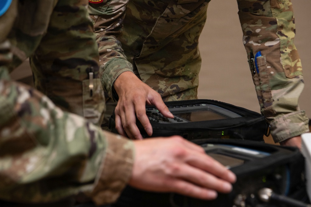 Two military personnel in OCP (Operational Camouflage Pattern) uniforms are handling what appears to be a ruggedized military electronic device or portable computing/communications equipment in a protective case. The image is a close-up shot focusing on their hands and the equipment. At least one patch is visible on the upper arm of one soldier.