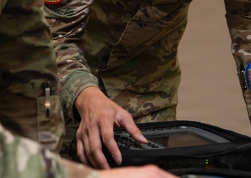 Two military personnel in OCP (Operational Camouflage Pattern) uniforms are handling what appears to be a ruggedized military electronic device or portable computing/communications equipment in a protective case. The image is a close-up shot focusing on their hands and the equipment. At least one patch is visible on the upper arm of one soldier.