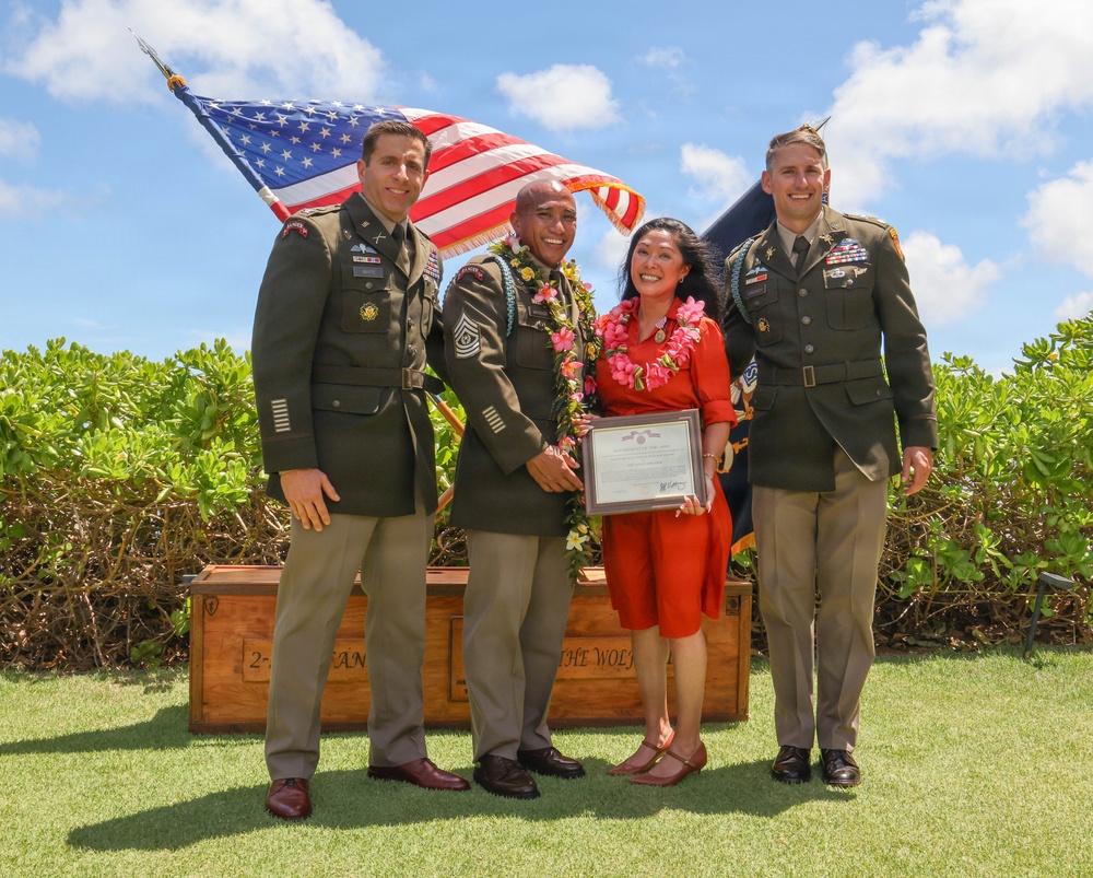 Four individuals pose for a formal military ceremony photograph outdoors, with an American flag waving behind them. Two uniformed U.S. Army soldiers flank a central male soldier wearing a floral lei and a civilian woman in a red dress, both also wearing leis, who is holding a framed certificate or award. The setting appears to be in Hawaii based on the tropical vegetation, leis, and outdoor ceremony setup.