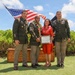 Four individuals pose for a formal military ceremony photograph outdoors, with an American flag waving behind them. Two uniformed U.S. Army soldiers flank a central male soldier wearing a floral lei and a civilian woman in a red dress, both also wearing leis, who is holding a framed certificate or award. The setting appears to be in Hawaii based on the tropical vegetation, leis, and outdoor ceremony setup.