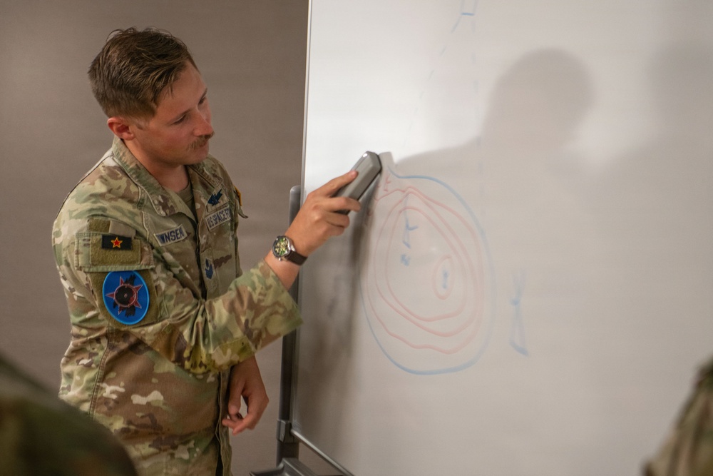 A U.S. Air Force service member in OCP (Operational Camouflage Pattern) uniform is presenting at a whiteboard, holding an eraser. The whiteboard displays concentric circles drawn in red and blue marker, suggesting a tactical or operational briefing diagram. The individual appears to be in a classroom or briefing room setting.