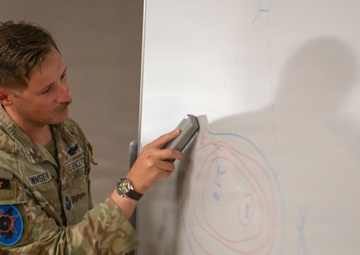 A U.S. Air Force service member in OCP (Operational Camouflage Pattern) uniform is presenting at a whiteboard, holding an eraser. The whiteboard displays concentric circles drawn in red and blue marker, suggesting a tactical or operational briefing diagram. The individual appears to be in a classroom or briefing room setting.