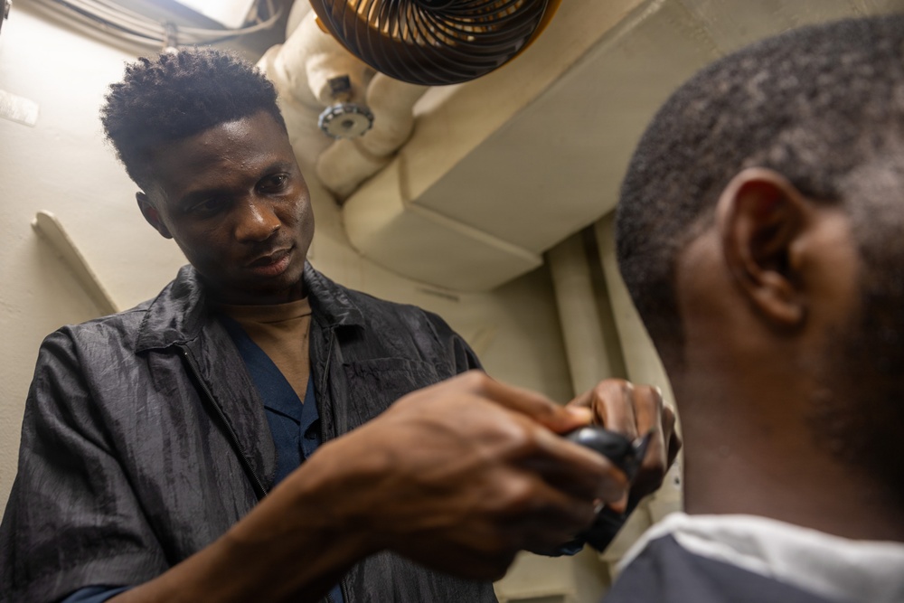 A man is cutting or grooming another man's hair using electric clippers in what appears to be a small, enclosed indoor space. The barber is focused and leaning in closely while working on the client, who is seated and wearing a white cape or collar. The setting appears informal, possibly a makeshift barbershop or small room.