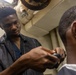A man is cutting or grooming another man's hair using electric clippers in what appears to be a small, enclosed indoor space. The barber is focused and leaning in closely while working on the client, who is seated and wearing a white cape or collar. The setting appears informal, possibly a makeshift barbershop or small room.